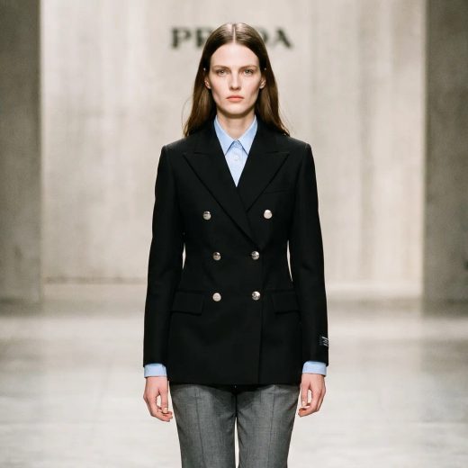 Женщина стоит на подиуме в стильном образе от Prada: строгий черный двубортный пиджак с серебристыми пуговицами, под ним светло-голубая классическая рубашка и серые классические брюки. Фон минималистичный, мягкое освещение, средний план, прямой взгляд.