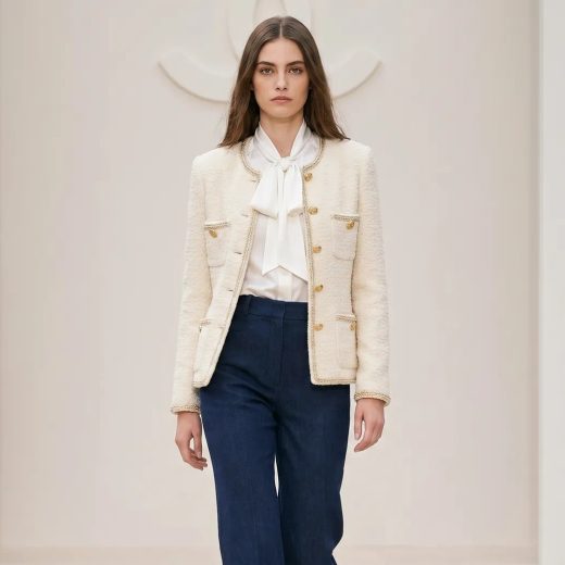 Женщина на подиуме в образе от Chanel: короткий жакет кремового оттенка из твида с золотыми пуговицами, лаконичная белая блуза и темно-синие прямые брюки с высокой талией. Освещение мягкое, фон минималистичный, средний план, прямой взгляд.