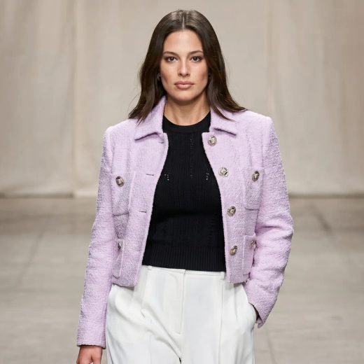 Женщина позирует на подиуме, на ней костюм от Versace: короткий светло-сиреневый жакет с фактурными пуговицами, под ним черный трикотажный топ и широкие белые брюки. Фон нейтральный, освещение мягкое, средний план, уверенный взгляд в камеру.